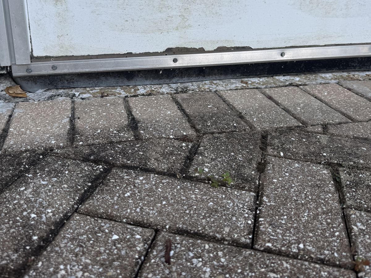 The bottom of an exterior door above a paver step, with a visible gap under a worn bottom seal.