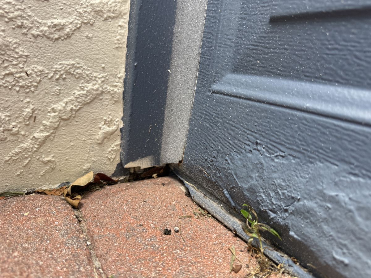 The bottom corner of a gray garage door, where the door seal stops short of the jamb and leaves a gap at the ground.