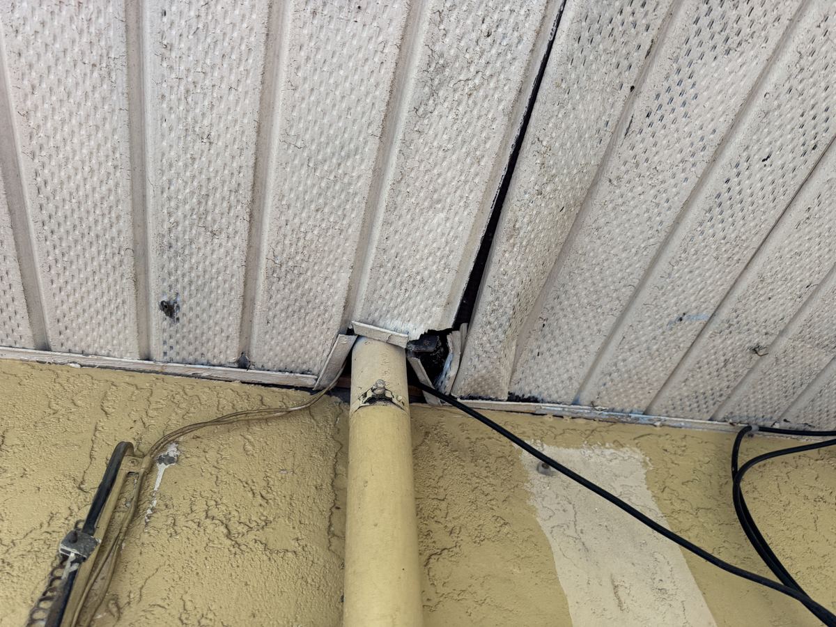 A pipe running up into a vented soffit, with a broken, open gap in the soffit panel beside it.